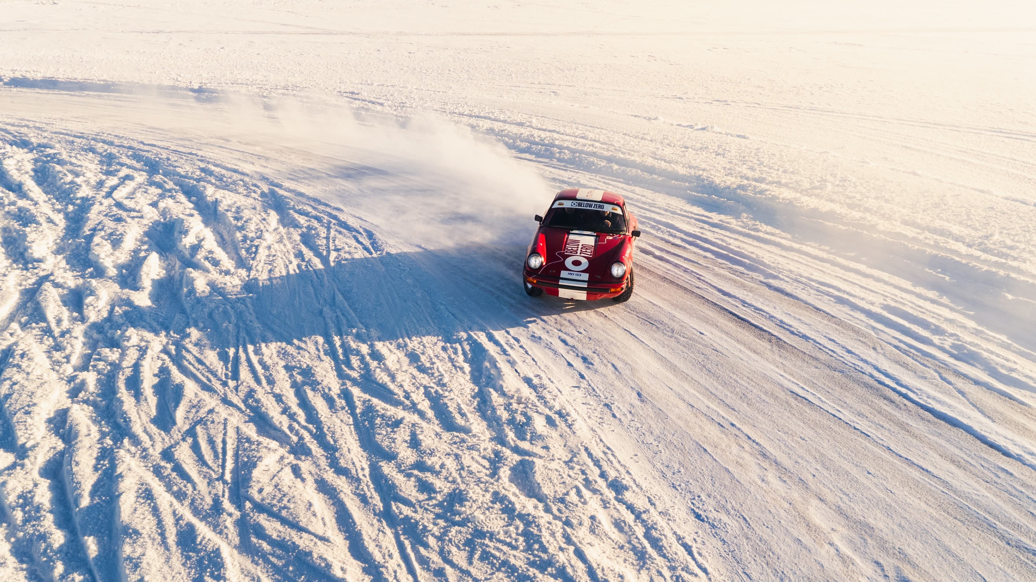 Red Porsche 911 rally car drifts on a snowy road, leaving tire tracks and a trail of snow dust under bright, clear skies.