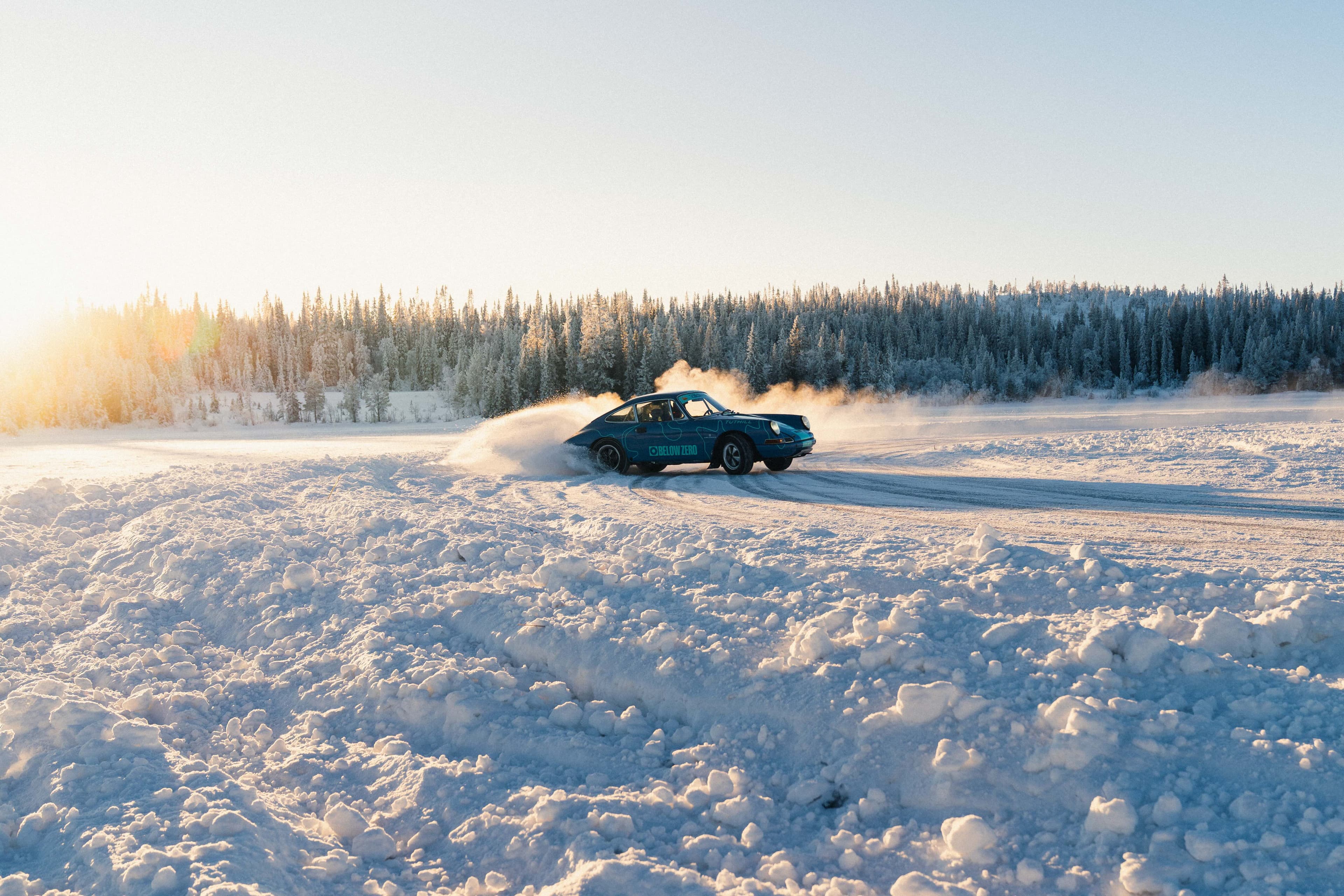 Porsche 911 rally car drifting on a snowy landscape at sunset, with a forest in the background. Snow billows behind the vehicle as it moves.