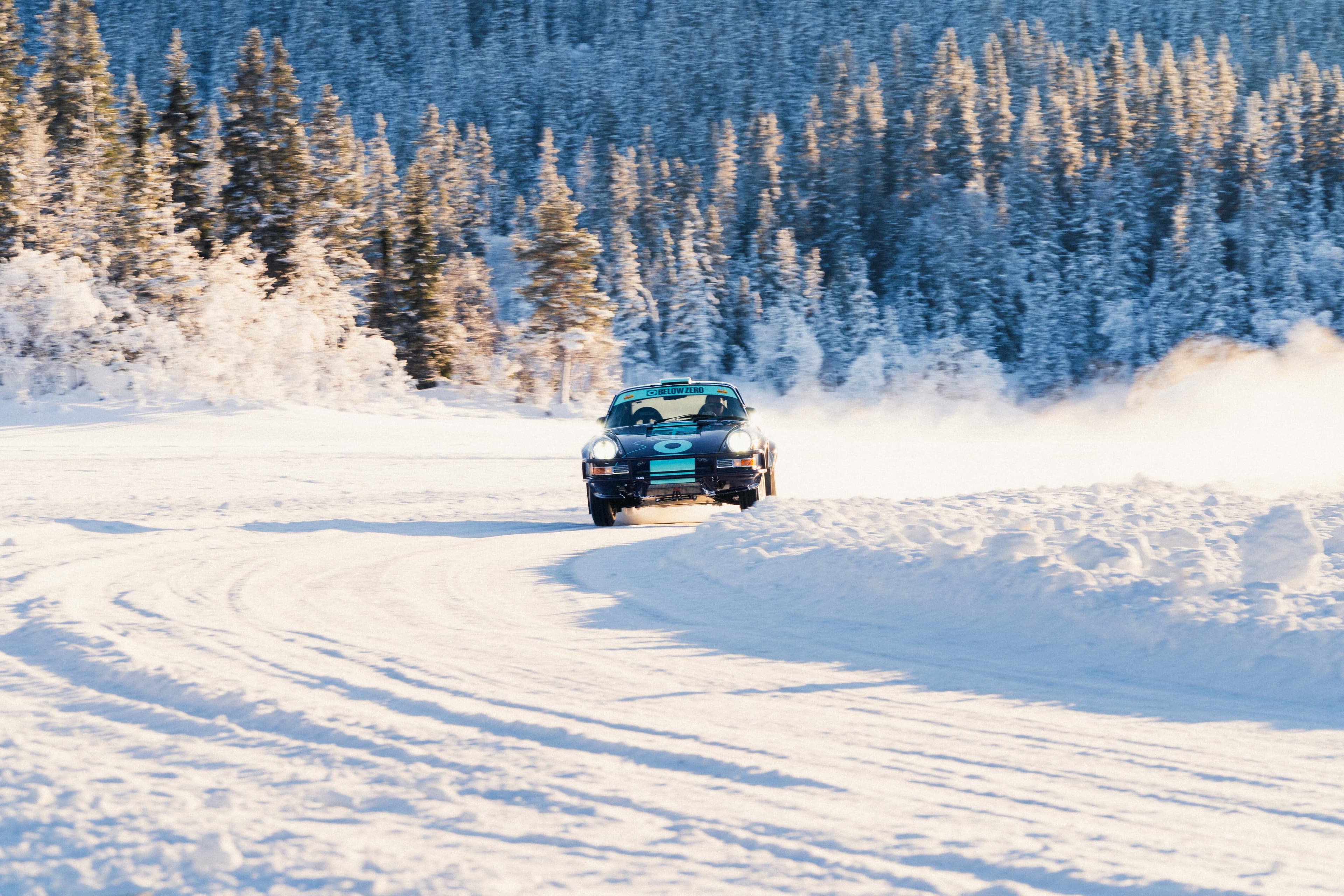 Porsche 911 rally car drifting on a snowy road surrounded by snow-covered trees and a forested landscape under a clear sky.